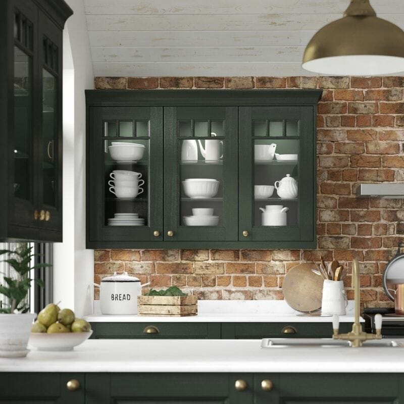 A cozy kitchen with green cabinets and a brick backsplash. The upper cabinets display white dishes. Theres a marble countertop with a bread box, fruit bowl, and kitchen utensils. A pendant light hangs from the ceiling.